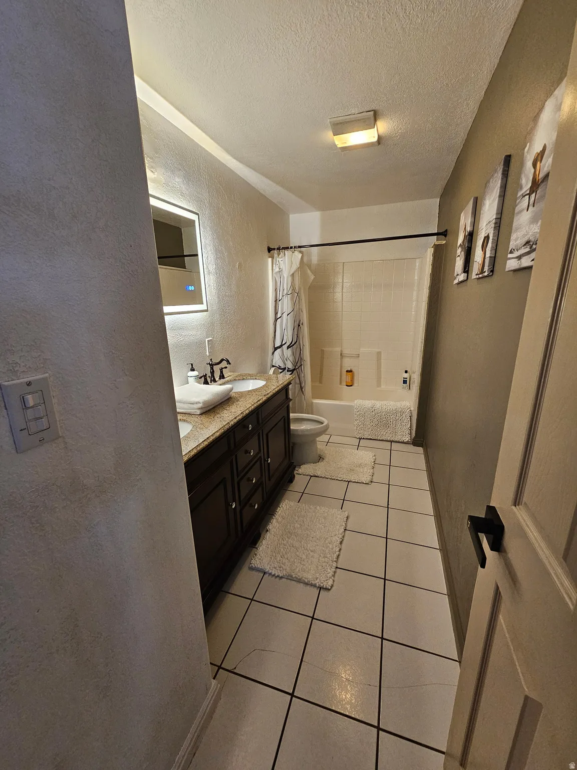 Bathroom with a textured wall, shower / tub combo, double vanity, a textured ceiling, and light tile patterned floors