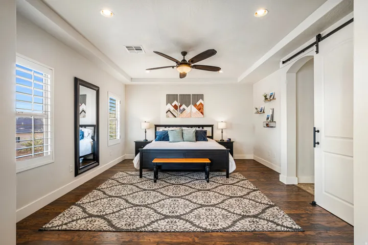 Bedroom with a barn door, a raised ceiling, dark wood-type flooring, a ceiling fan, and recessed lighting