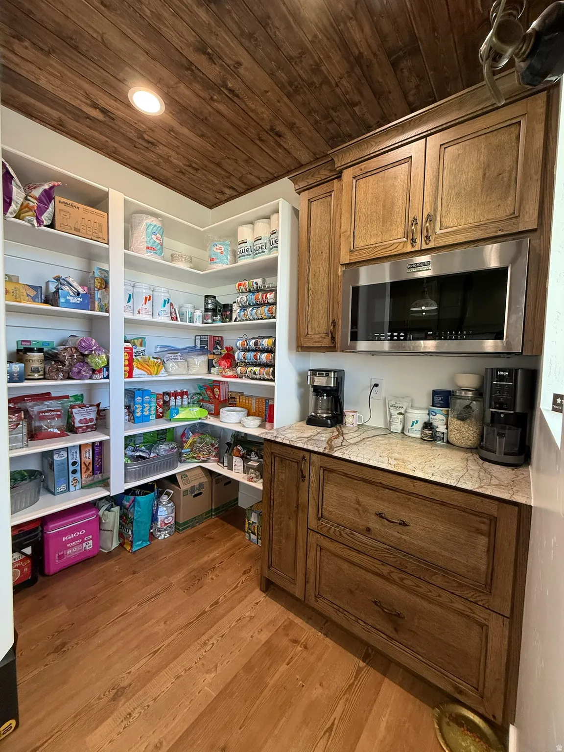 Spacious pantry featuring extensive built-in shelving, wood-finish flooring, a wood plank ceiling, custom wood cabinetry, and a stainless steel microwave