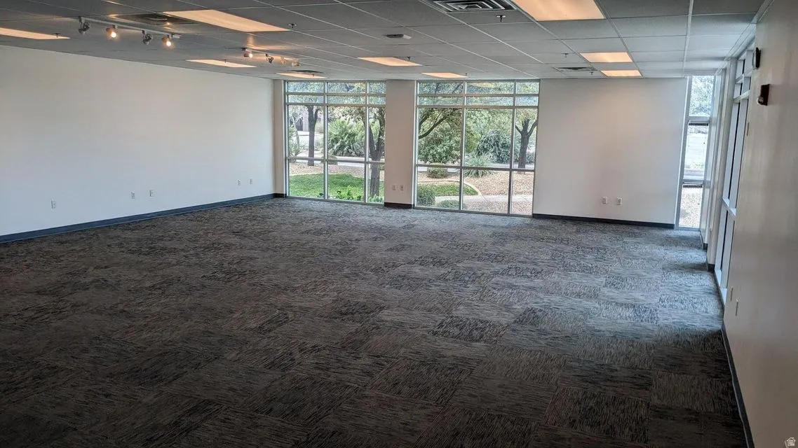 Empty room with dark carpet, track lighting, a paneled ceiling, and expansive windows