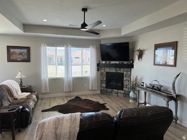 Living area with a tray ceiling, ceiling fan, wood finished floors, a textured ceiling, and a stone fireplace