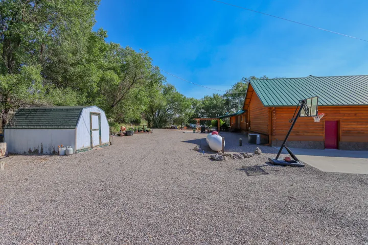 Expansive gravel parking area featuring a storage shed with a green shingle roof, a log-sided structure with a green metal roof, a covered patio, and a concrete pad