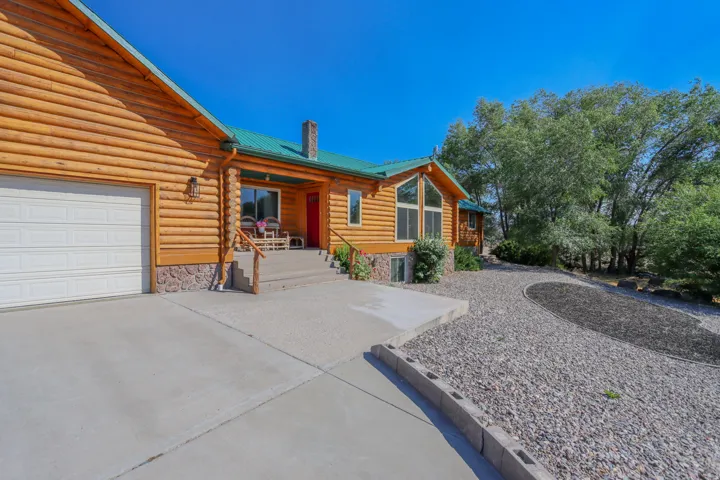 Log-style exterior featuring a green metal roof, attached garage, stone foundation accents, a covered front porch, and a gravel driveway