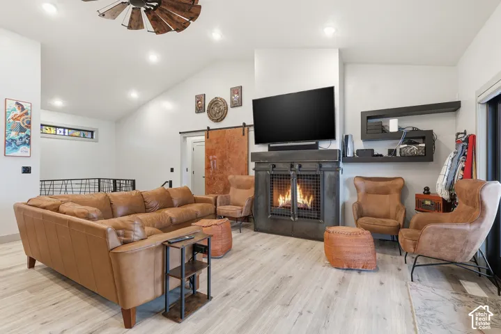 Great room featuring ceiling fan, light wood-style flooring, a warm lit fireplace, lofted ceiling, and a barn door