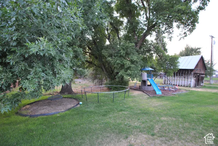 Expansive lawn area featuring mature trees, a designated play structure with a slide, and a rustic storage building with a green roof