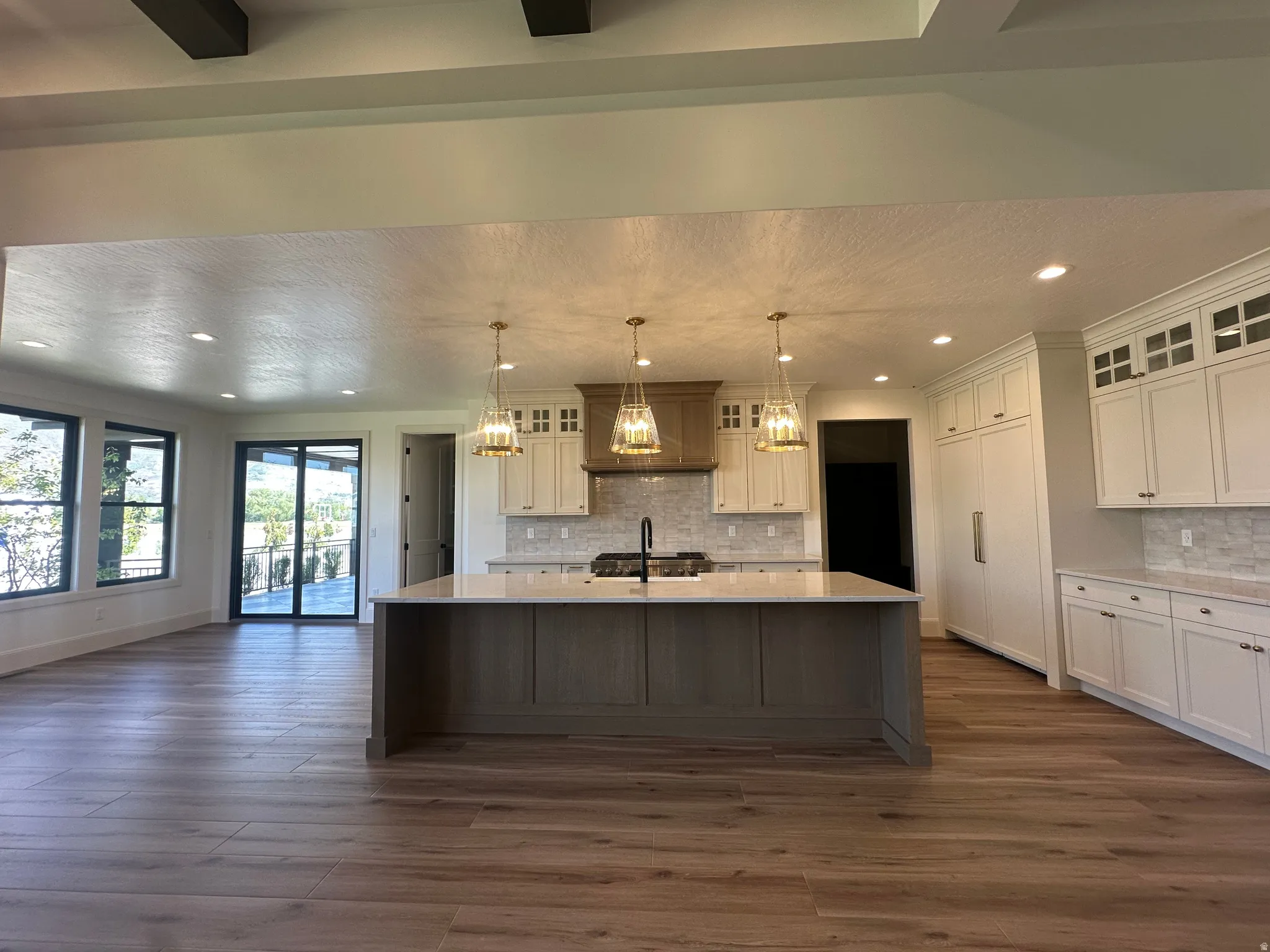 Open-concept living space featuring wood-finish flooring, a kitchen island with pendant lighting, white cabinetry, integrated appliances, and large windows with exterior access