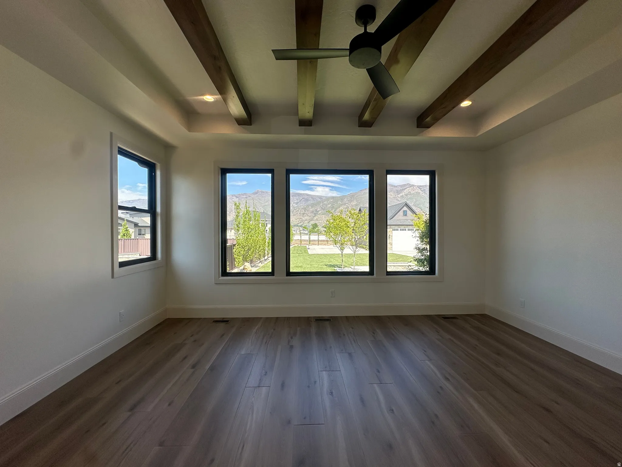 Spacious primary bedroom featuring wood-finish flooring, a tray ceiling with stained wood beams, recessed lighting, and a ceiling fan