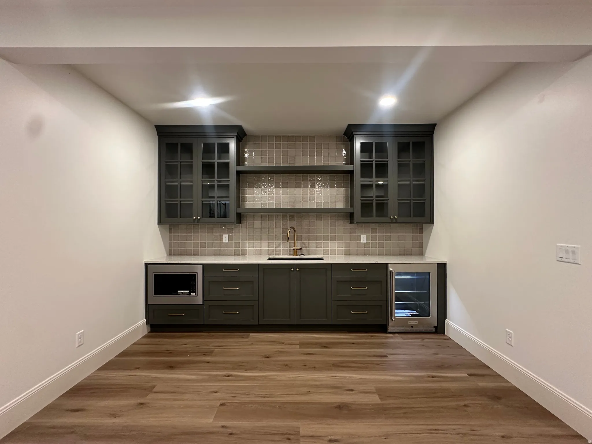 Built-in wet bar featuring dark gray cabinetry, glass-front upper cabinets, open shelving, a tiled backsplash, and a beverage cooler