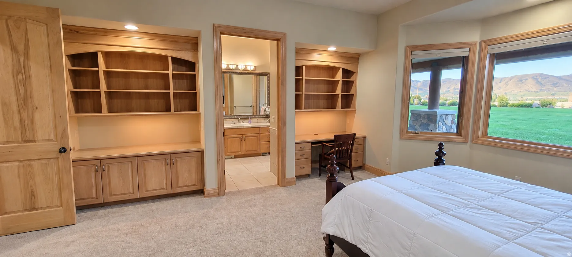 Built-in shelving units, integrated desk with task lighting, dual windows with wood trim, wall-to-wall carpeting, and an adjacent bathroom with a double vanity
