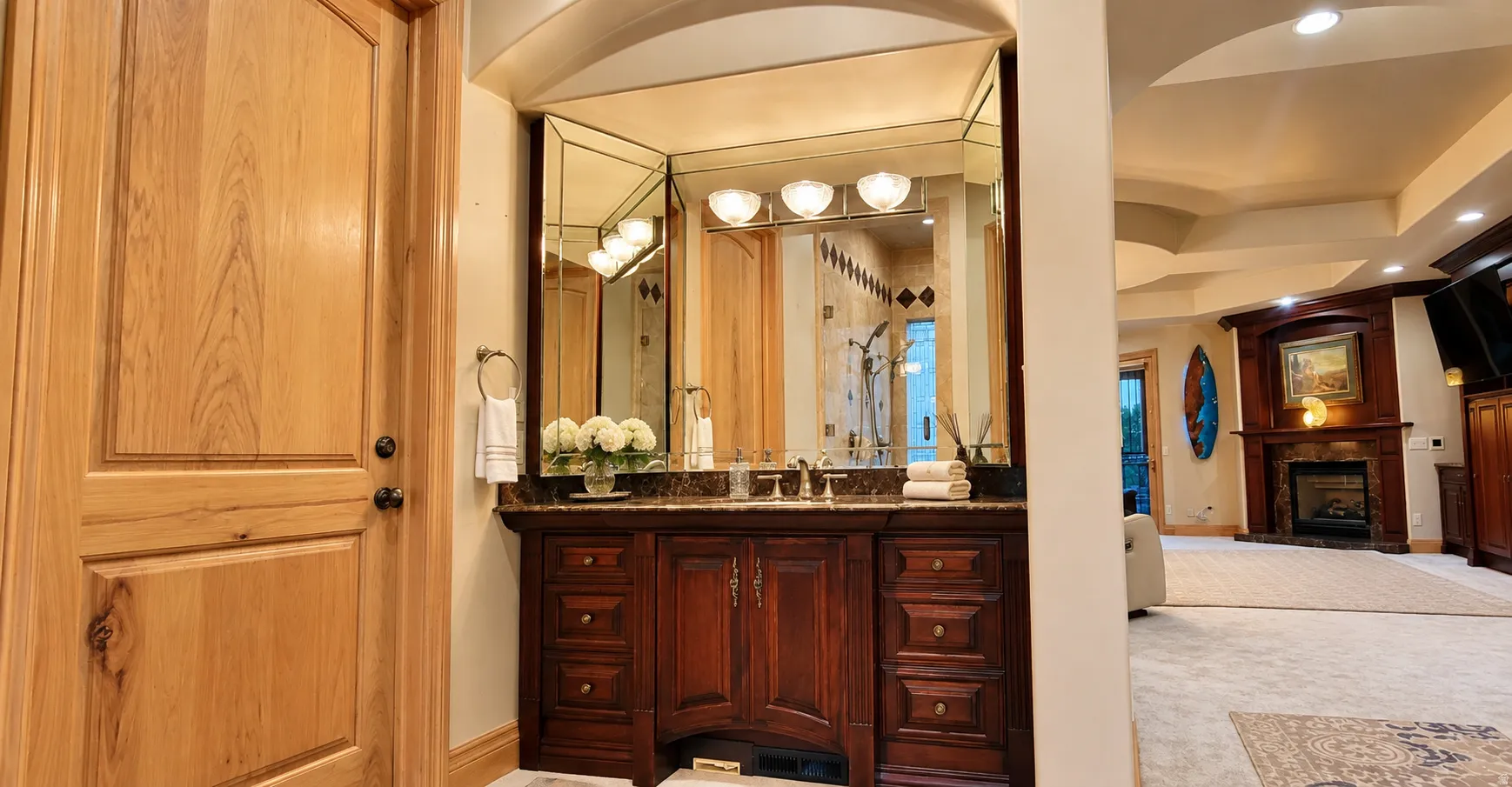 Hardwood paneled door with dark bronze hardware, wood-finish vanity with stone countertop, beveled mirror, and wall-mounted light fixtures