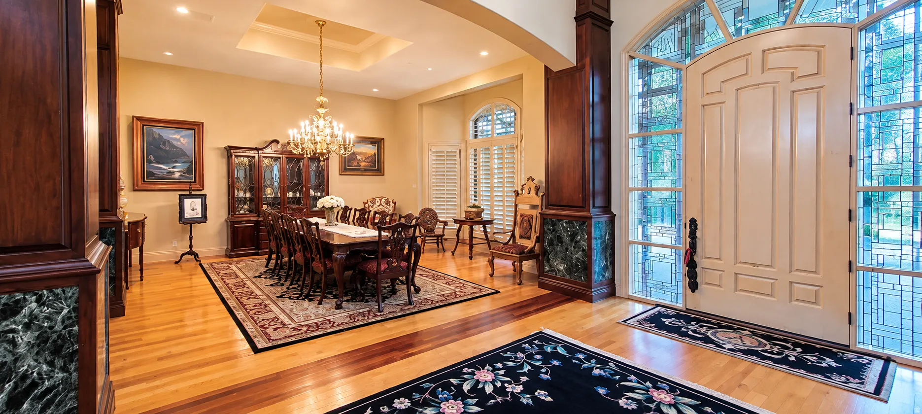 Hardwood flooring with contrasting inlay, tray ceiling with chandelier, arched windows with plantation shutters, decorative columns with marble accents, and an ornate front entry door with leaded glass sidelights