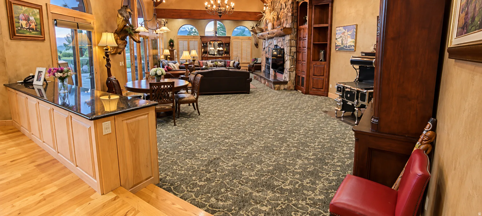 Hardwood flooring, kitchen island with dark countertop, multiple arched windows, stone-clad fireplace, and an ornate chandelier