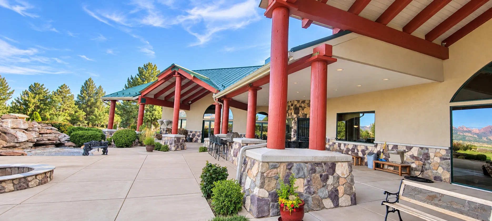 Expansive patio with stone-accented fire pit, paved hardscaping, natural rock landscaping, and a covered walkway supported by robust red columns