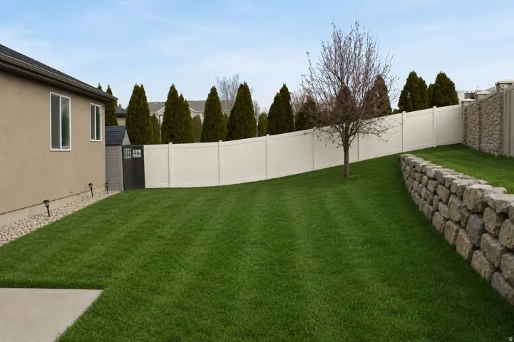 Expansive lawn featuring a vinyl privacy fence, a stone retaining wall, and a concrete patio section.  AI Adjusted Photo
