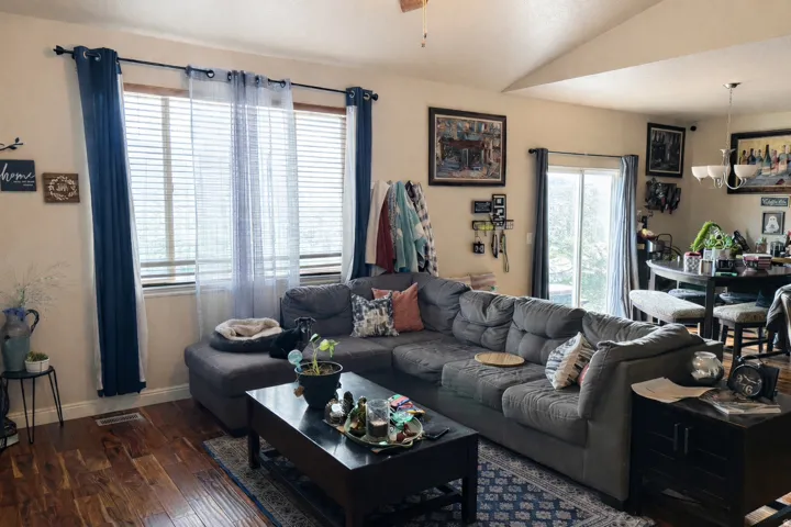 Vaulted ceiling living area featuring wood-finish flooring, a large window with blinds, and a sliding glass door