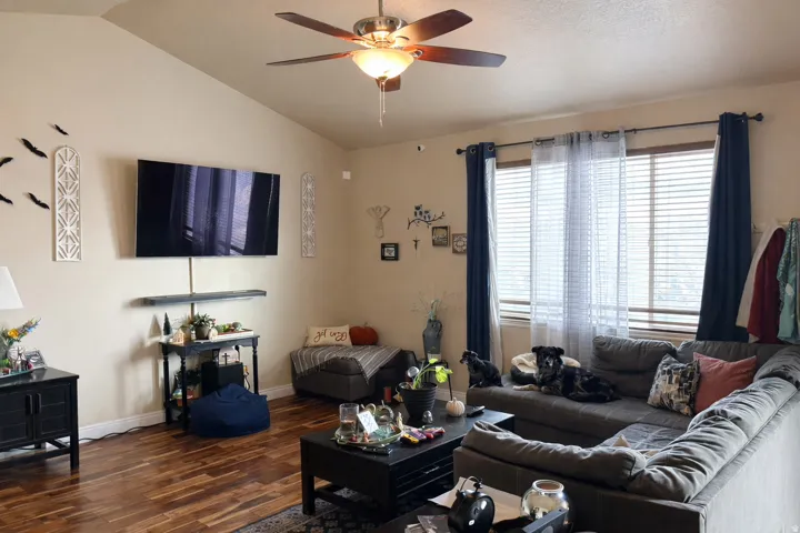 Vaulted ceiling living space featuring wood-finish flooring, a contemporary ceiling fan with integrated lighting, and multiple windows with blinds