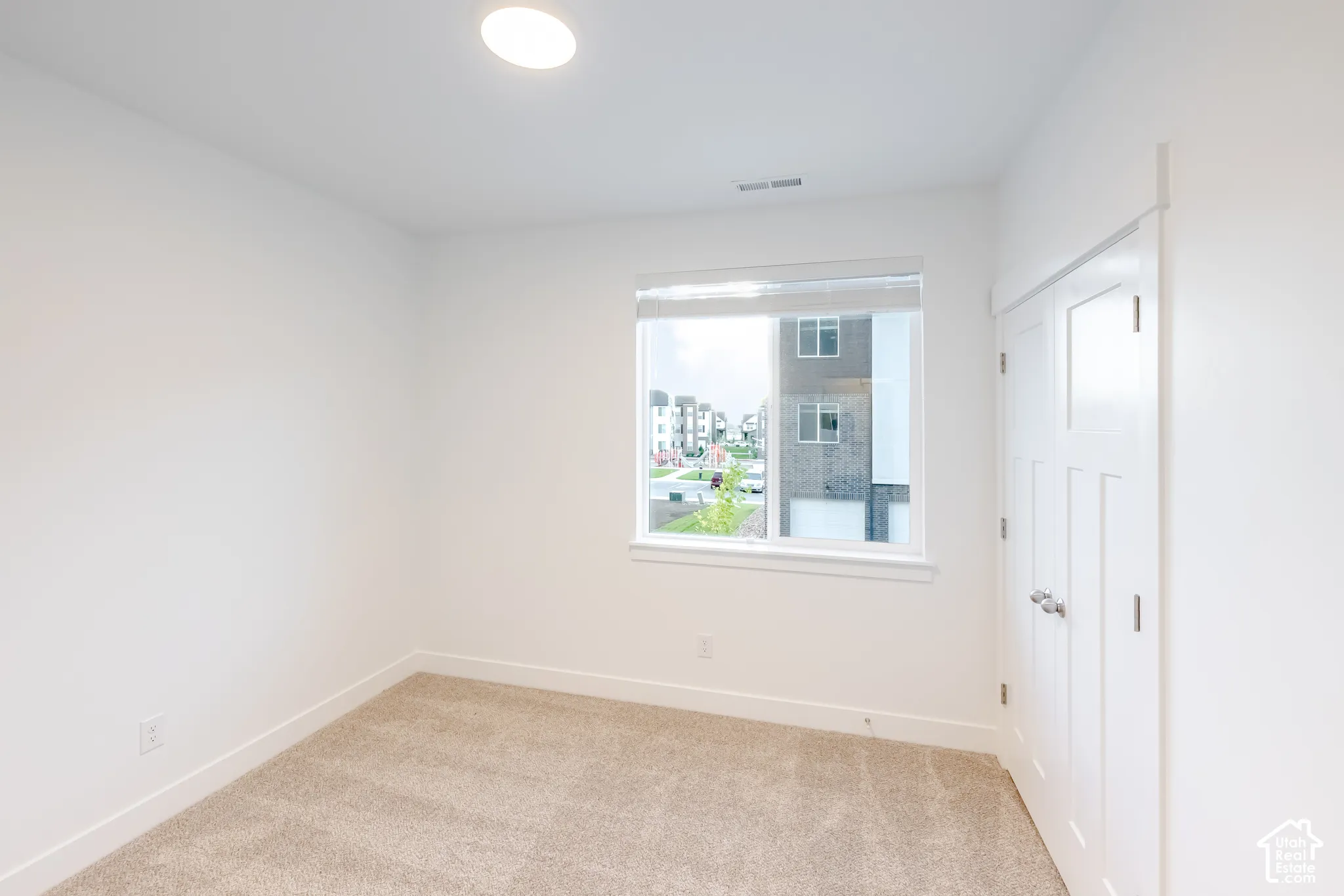 Neutral-toned wall-to-wall carpeting, single-pane window with white roller shade, and double-door closet with satin-nickel hardware