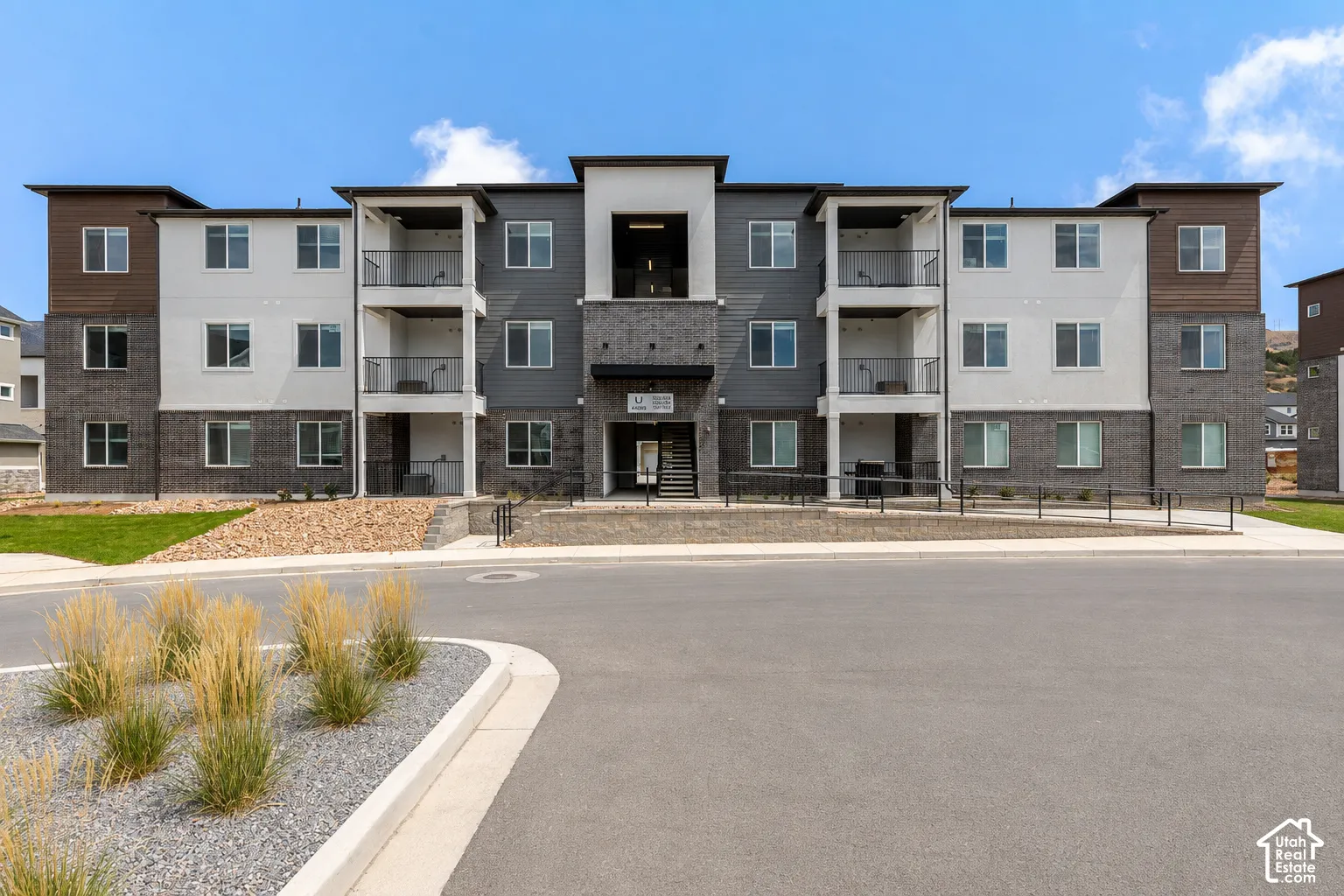 Multi-story residential exterior featuring a mixed-material facade of stone veneer, stucco, and wood-finish siding, complemented by private balconies and paved ground-level entry