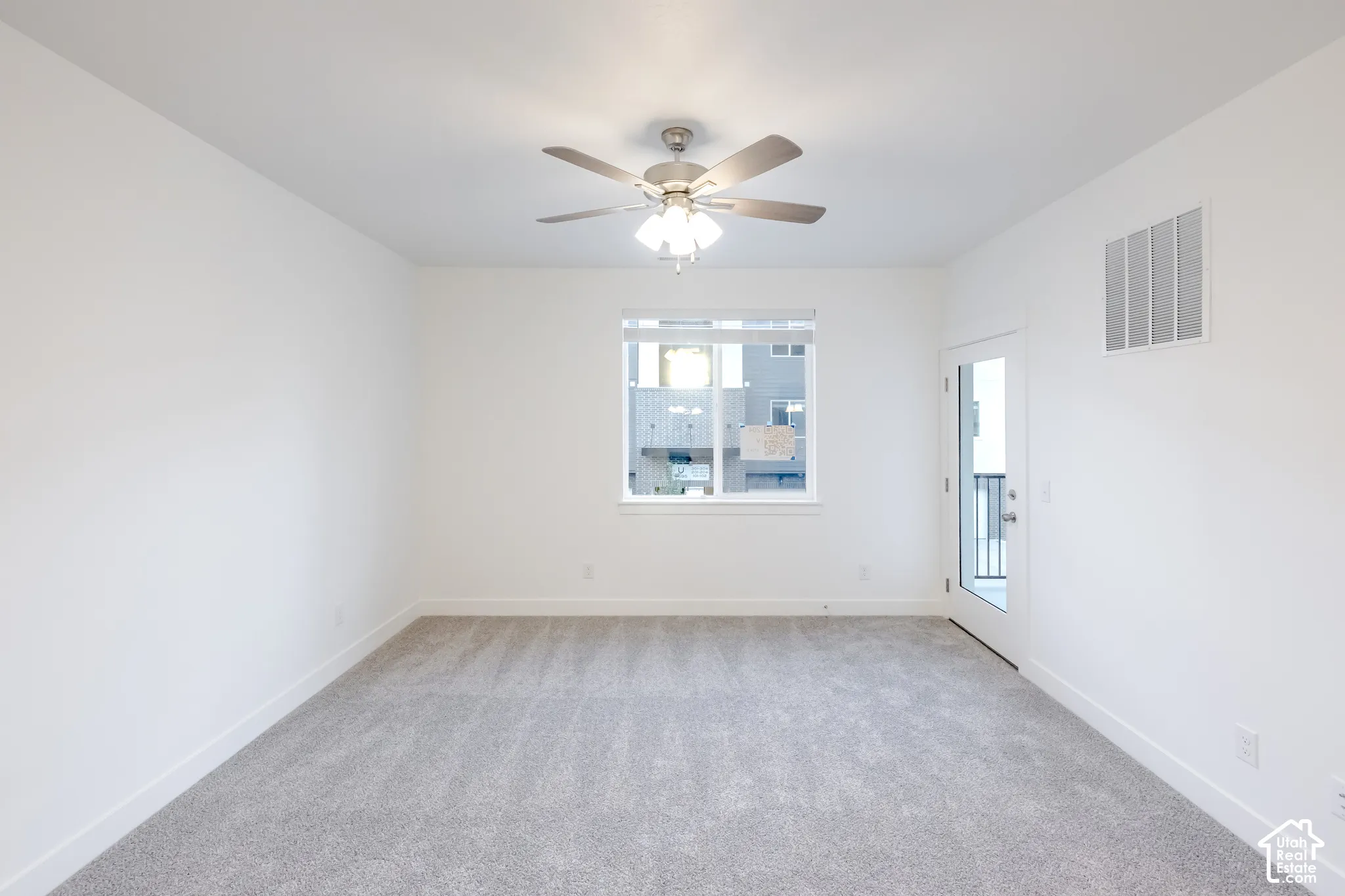 Neutral-toned wall-to-wall carpeting, white walls, multi-blade ceiling fan with integrated lighting, window with horizontal blinds, and a door providing exterior access
