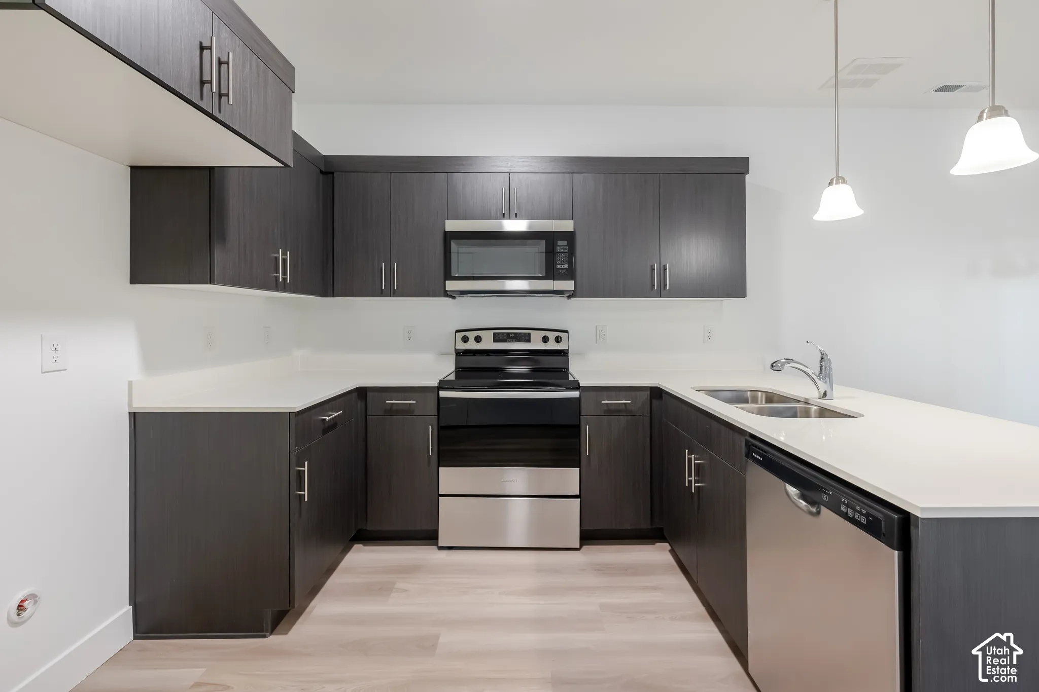 Kitchen featuring dark-finish cabinetry, stone-composite countertops, stainless steel appliances, light-toned wood-finish flooring, and dual hanging pendant lights