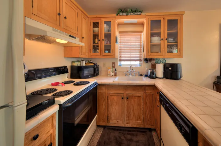 Apartment kitchen with white appliances, tile counters, wood finish cabinets, a peninsula, and glass fronted cabinets