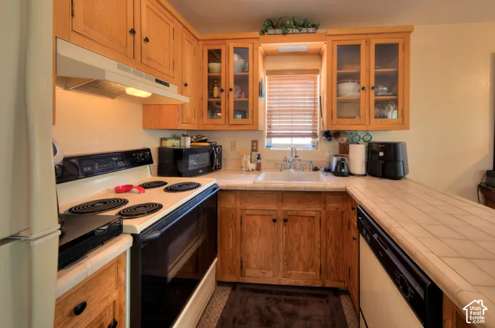 ADU Kitchen with white appliances including a dishwasher,  tile counters, glass front cabinets and a peninsula offering plenty of counter space.