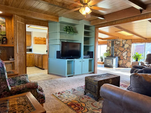 Living room with a wood ceiling with exposed beams, a wood stove, ceiling fan, and light colored carpet