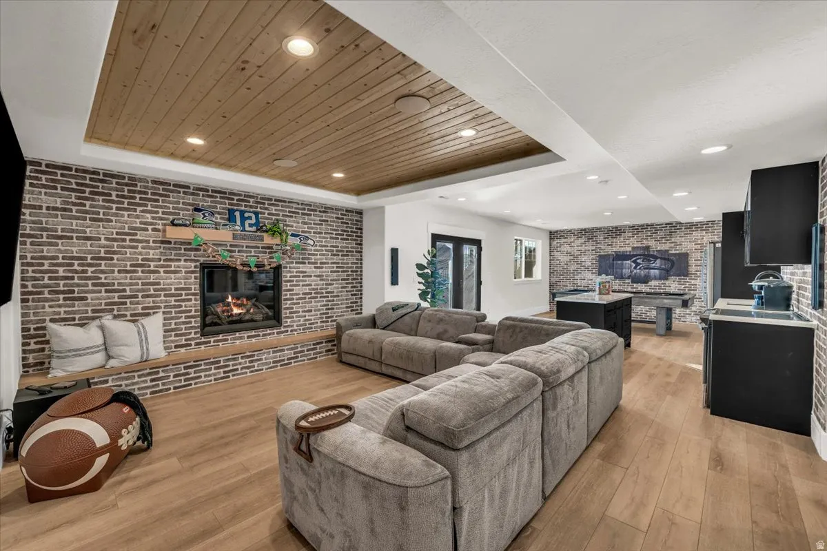 Living area with brick wall, a fireplace, light wood-style flooring, a wooden tray ceiling, and recessed lighting