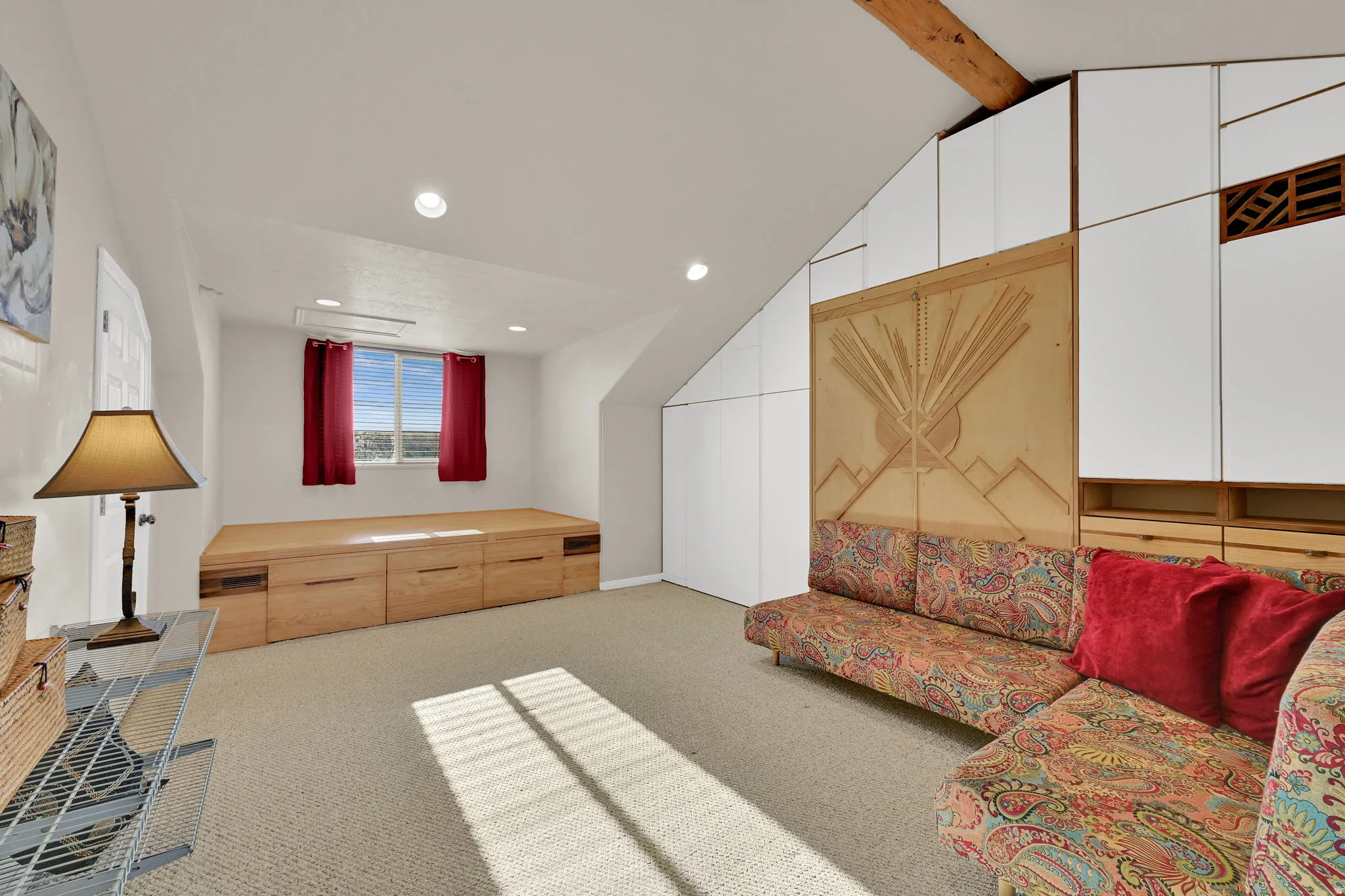 Carpeted room featuring a built-in wooden platform with integrated drawers, recessed lighting, and a window with red drapes