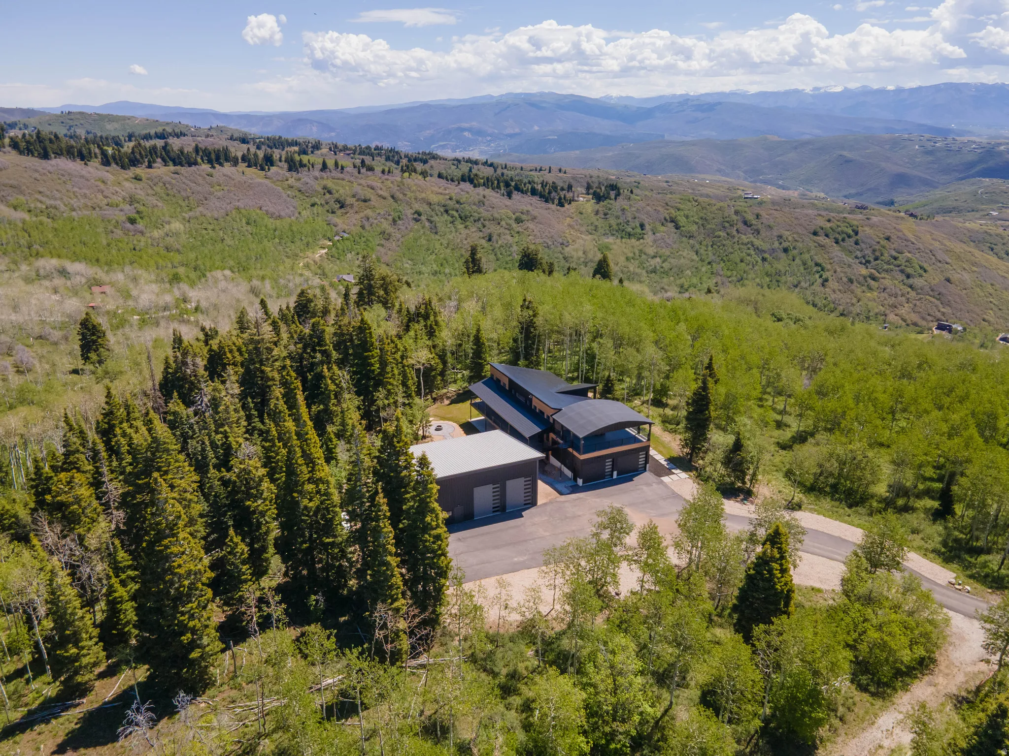 Modern architectural property with dark roofing, multiple balconies, a gravel driveway, and a separate structure with a metal roof, all set within a forested, mountainous landscape