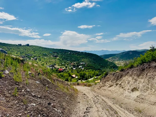 Expansive mountain landscape featuring a dirt road, surrounding hillsides with natural foliage, and distant residential structures