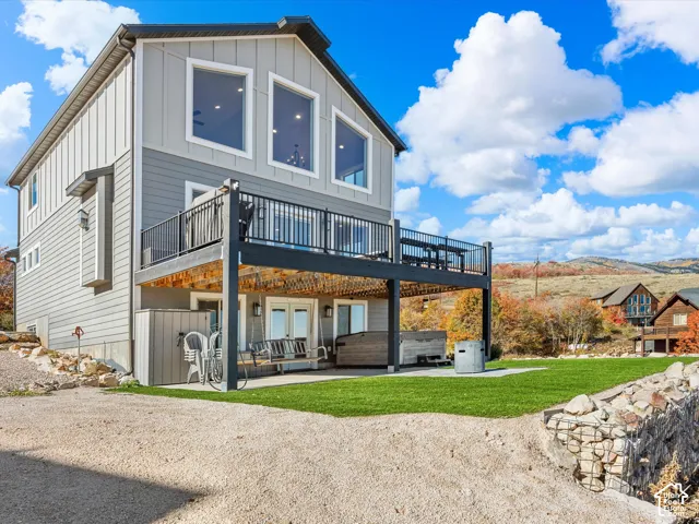 Exterior siding in varying grey tones, large deck with black railings, lower-level patio with double doors, manicured lawn area, and a gravel and asphalt drive