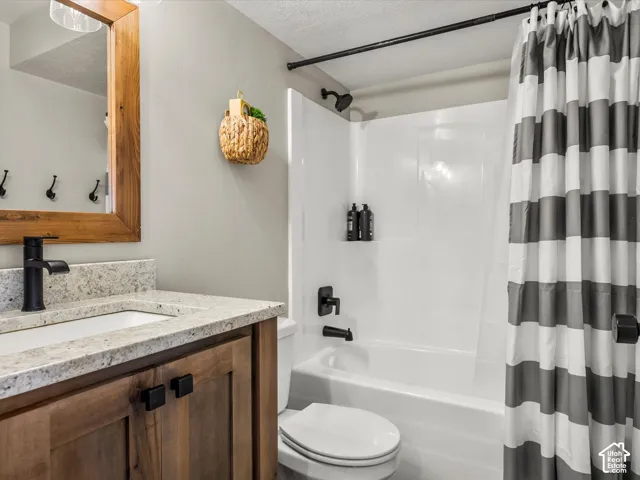 White bathtub with integrated shower, single vanity with wood-finish cabinetry, light-toned countertop, and matte black fixtures