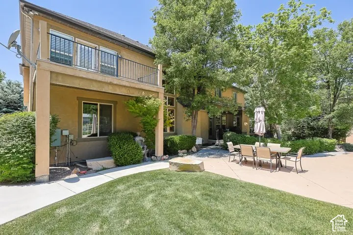 Two-story stucco exterior with a second-level balcony, ground-level patio, mature trees, and a grass lawn