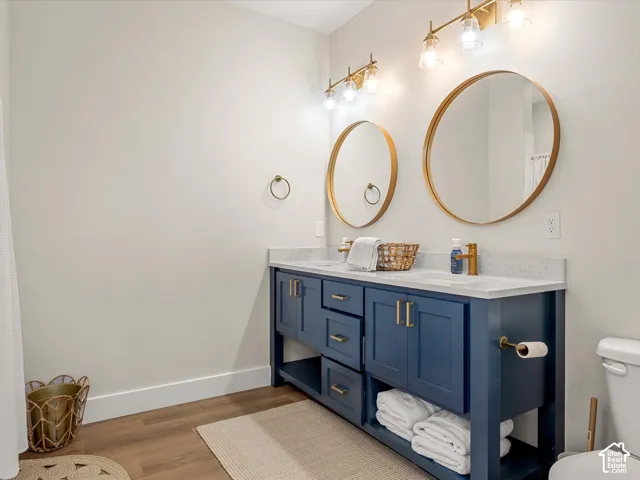 Dual round mirrors with brass frames, dual brass vanity lighting, a blue double vanity with gold hardware, a white countertop, and wood-finish flooring