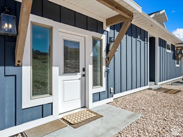 Board and batten siding with white trim, a white door with an integrated window, a concrete entry, and decorative wood beams