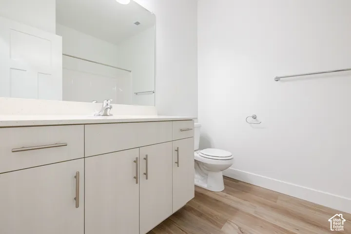 Bathroom featuring light wood-finish cabinetry, a solid surface countertop, an undermount sink with a polished chrome faucet, a full-width wall mirror, and wood-finish flooring