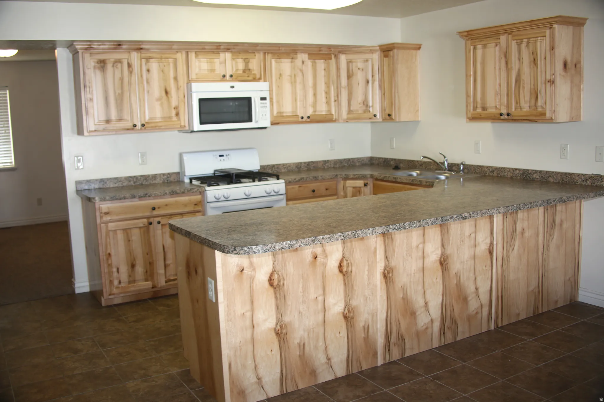 Kitchen with white appliances, a peninsula, light wood finish cabinets, and dark countertops