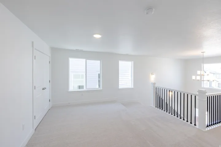 Carpeted loft area featuring white walls, two windows, a white railing with black spindles, and a multi-light chandelier