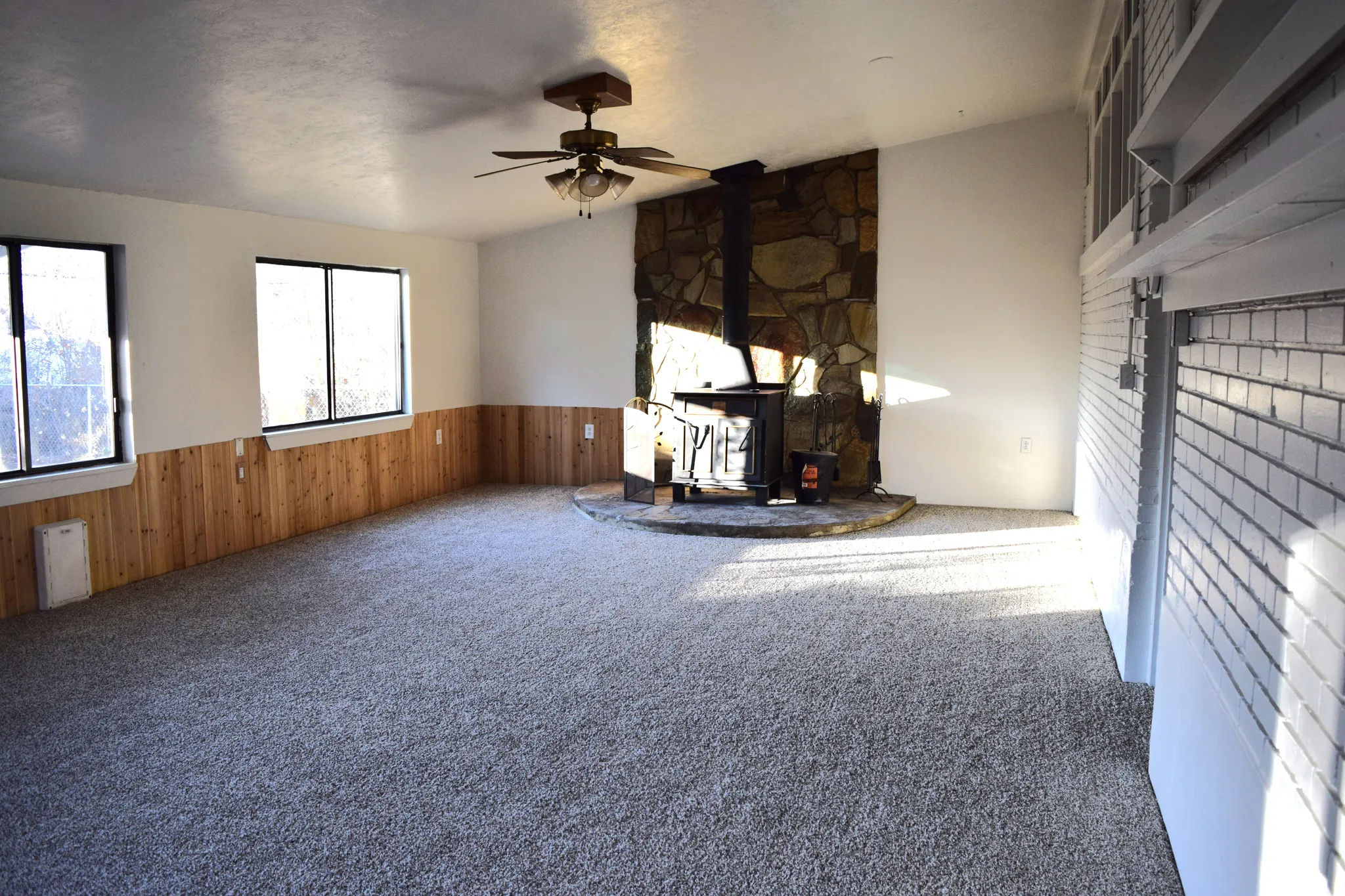 Unfurnished living room featuring a wood stove, wooden walls, carpet floors, wainscoting, and a ceiling fan