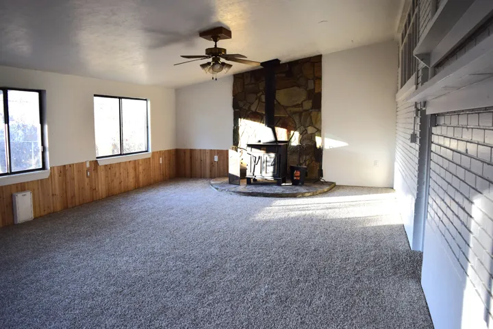 Unfurnished living room featuring a wood stove, wooden walls, carpet floors, wainscoting, and a ceiling fan