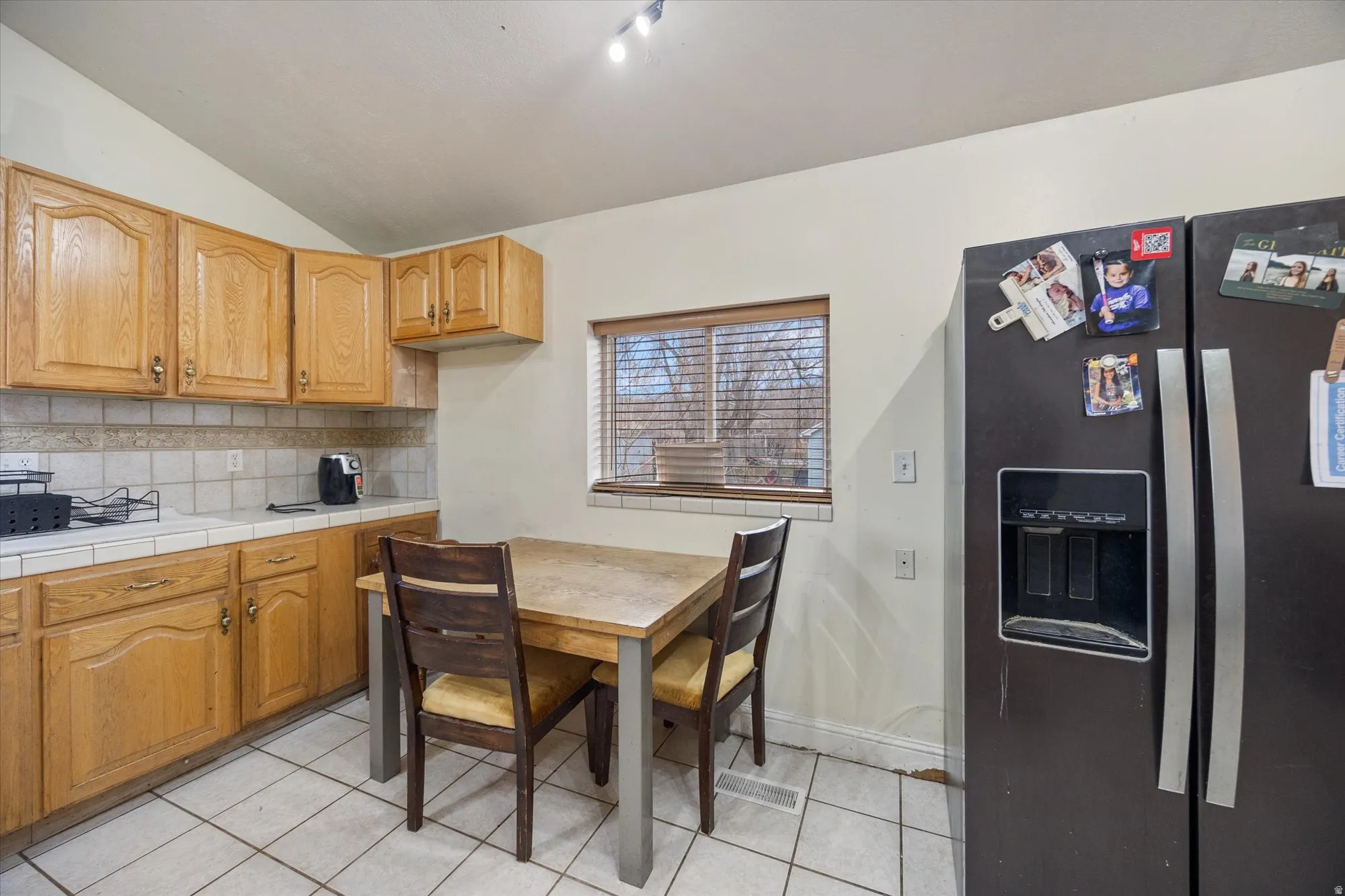 Kitchen featuring black refrigerator with ice dispenser, tile countertops, light tile patterned floors, backsplash, and vaulted ceiling