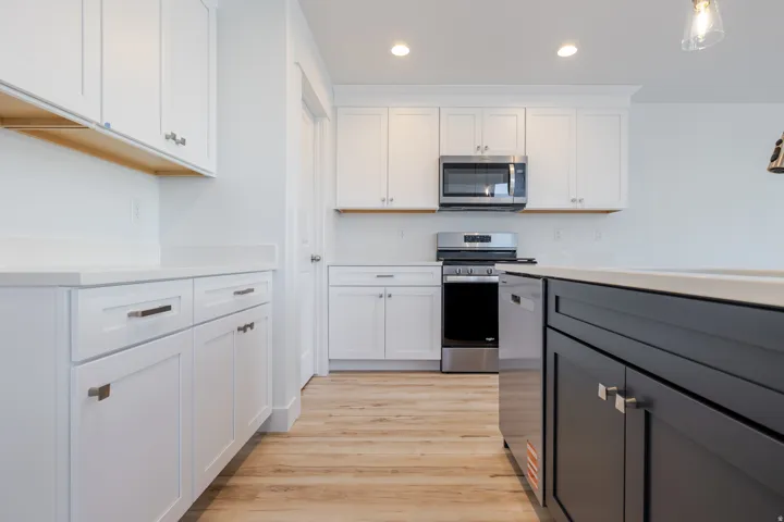 Modern kitchen featuring white shaker-style cabinetry, a contrasting dark-toned kitchen island, light-toned countertops, stainless steel appliances, and wood-finish flooring