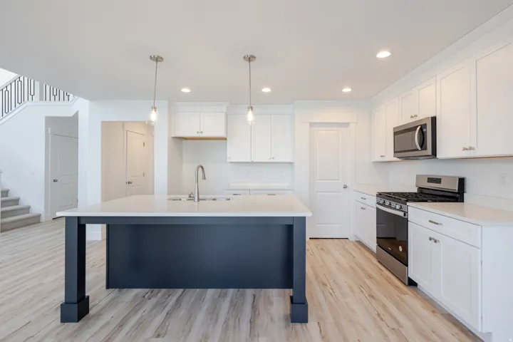 Kitchen featuring a dark-toned island with a white countertop and two pendant lights, stainless steel appliances, white cabinetry, and wood-finish flooring
