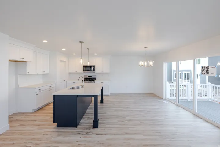 Open-concept living space featuring wood-finish flooring, a kitchen island with integrated sink, white shaker cabinetry, stainless steel appliances, and a sliding glass door opening to an outdoor deck