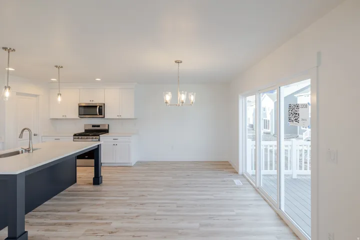 Contemporary kitchen and dining area featuring a dark blue island with integrated sink, stainless steel appliances, white cabinetry, light wood-finish flooring, and a sliding glass door opening to an outdoor deck