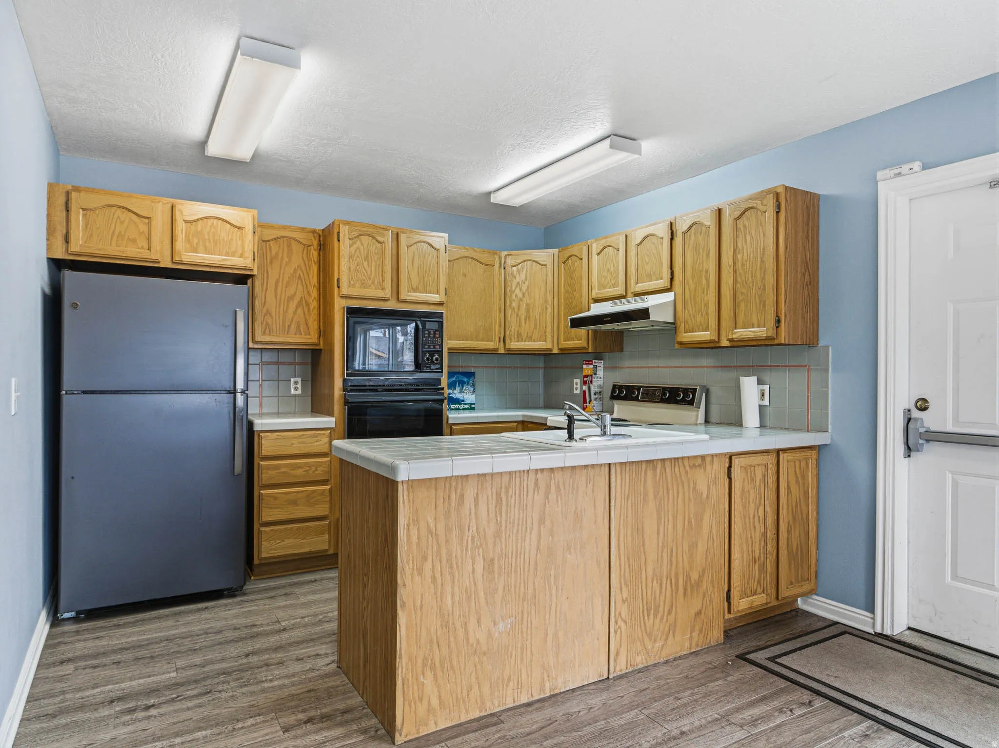 Kitchen featuring black appliances, a peninsula, tile counters, decorative backsplash, and wood finished floors