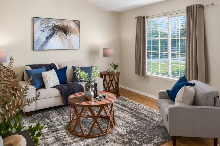 Spacious living area featuring light-toned walls, wood-finish flooring, and a large window with horizontal blinds