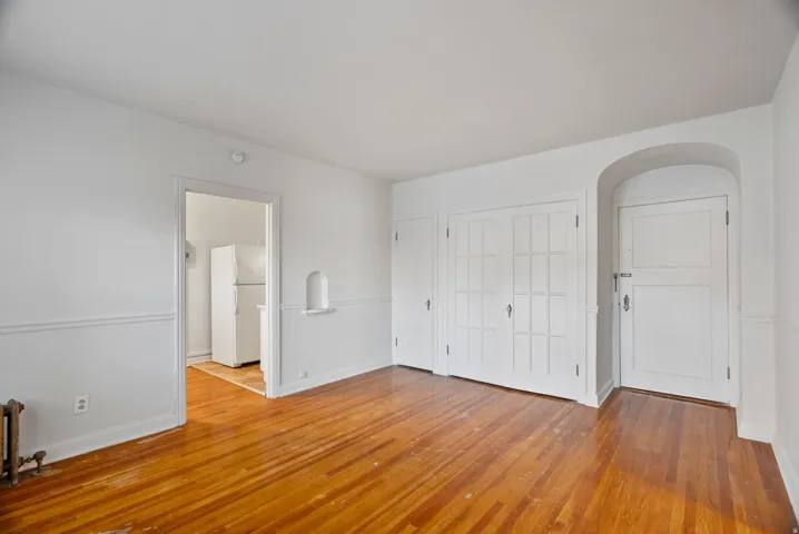 Unfurnished bedroom featuring freestanding refrigerator, light wood finished floors, radiator heating unit, and arched walkways