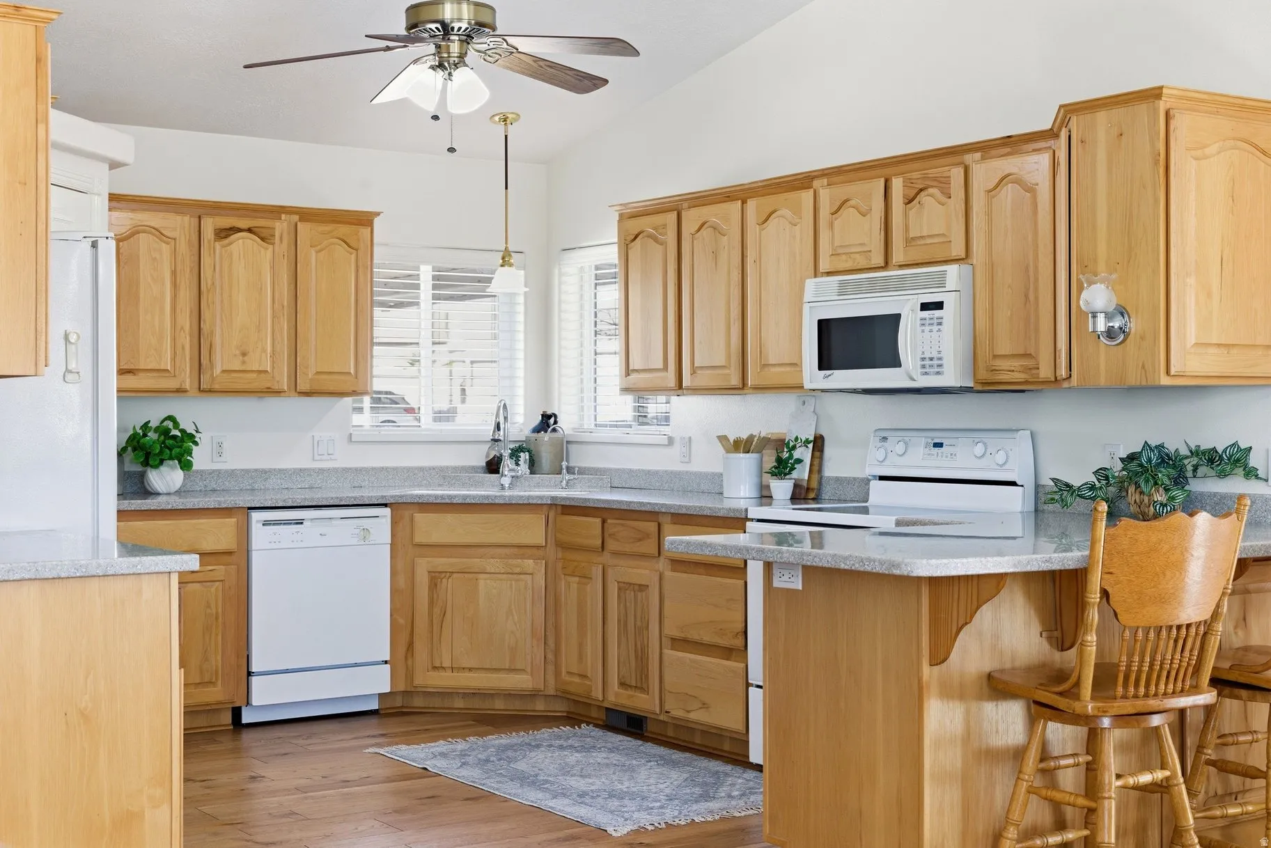 Kitchen featuring decorative light fixtures, ceiling fan, white appliances, light wood finish cabinets, and light stone counters